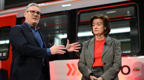Getty Images Sir Keir Starmer, a man in a navy suit with glasses and grey hair, stands next to Eluned Morgan, a woman in a grey suit with brown hair, in front of a stationary train.