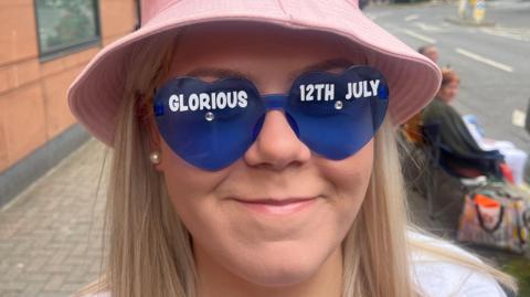 Woman with blond hair looks straight into camera smiling, wearing a pink hat and love heart shaped blue glasses with 'glorious 12th july' written across the frame in white