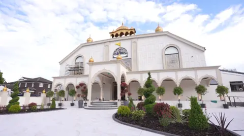 Guru Nanak Sikh Gurdwara The building is white with gold-looking domes on top. There are archways with trees in front and a wide, open path and entrance area.