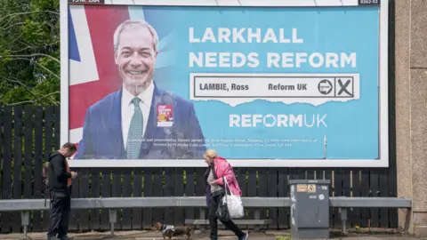 A large billboard image for reform featuring Nigel farage and a Union flag, two people walk in the foreground.