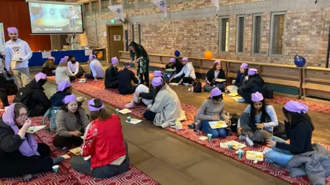 A group of people sitting down on red and white rugs on the floor of a chapel while eating and talking. They are all wearing purple headwear.