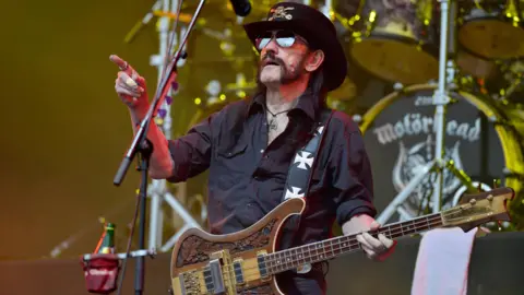 PA Media Motörhead frontman Lemmy, holding a bass guitar and holding his hand up pointing at something. He is wearing sunglasses, a black hat and a black shirt. He has a dark muttonchop-style beard. Behind him is a drum kit - the bass drum has a Motörhead logo on it.