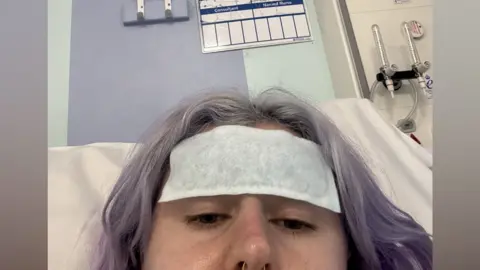 Abbie Filer A young woman with short, light purple hair lies in a hospital bed with a patch on her head.