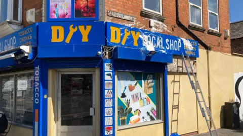 Alex Pope/BBC The outside of a shop front, with white paint on it. The shop front is most blue. There is a ladder up against the wall to the right and a door in the middle of the building. The walls are painted cream. 