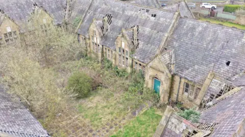 CAD Aerial A run-down building which is buil around a courtyard. It has a damaged roof and overgrowth across all the grounds. It is an aerial image showing the school building with a green door.