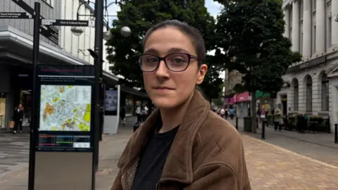Gemma Dillon/BBC Image shows a person wearing a black top and a brown jacket. They are wearing glasses and have their hair tied back in a ponytail. The image is of a street behind them.