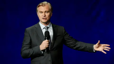 Reuters Christopher Nolan in a suit speaking on stage into a hand-held microphone with the other arm outstretched
