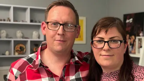 BBC Mike and Lisa Buller stand in their living room with shelves behind them and pictures and photos on the wall.