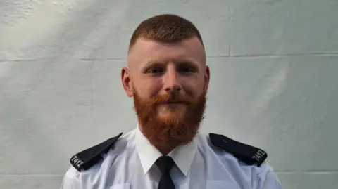 Charlie Jones has a ginger beard and short ginger hair. He wears a white and black police uniform. He looks at the camera, standing in front of a white wall.