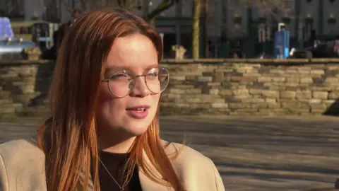 Margarida Vasconcelos with red hair, glasses, and a black t-shirt and beige coat, looks to the right of the camera, behind her is a brick wall 