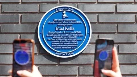 UMPF A blue circular plaque commemorating Brad Kella on a grey brick wall, with two mobile phones held up in front. It reads: This plaque commemorates the adjacent spot where, in August 2014, self-taught pianist Brad Kella first played at Liverpool ONE. It was the start of a personal journey that led to him winning series two of Channel 4's The Piano in 2024 and later achieving a number one debut album, Phobe's Melody. Liverpool One has celebrated local talent with its annual piano festival, Tickle the Ivories, since 2011.