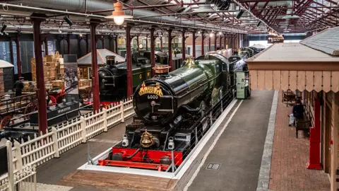 Swindon Borough Council Green and black steam trains in a museum. There stationary at platforms with people sitting at the side.