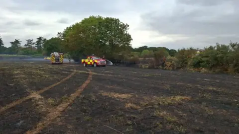 Fire engine and other fire service vehicles on a black charred field - firefighters are in the distance dousing a fire