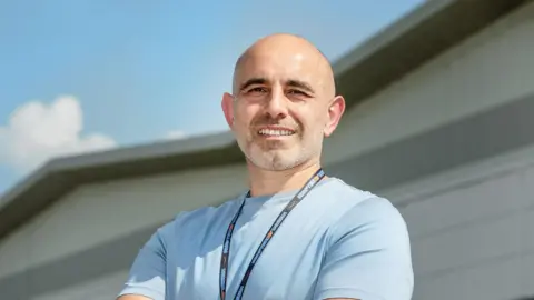 Amazon A man smiles at the camera. He wears a light blue t-shirt and a lanyard. In the background is a grey/white building. 