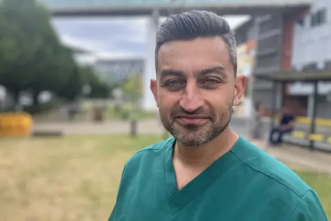 BBC/James Gallagher The image shows a close-up portrait of a person standing outdoors, wearing teal V-neck medical scrubs. The photograph is sharply focused on the person in the foreground, while the background is blurred with a shallow depth-of-field effect. In the background, there appears to be a grassy area, trees, a modern building, and a bus stop or shelter with a few people nearby. 