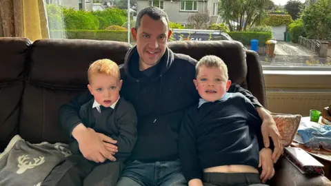 Lorraine Wallace A person in a black hoodie sits on a brown leather sofa between two young children wearing school uniforms. The three are positioned in front of a large picture window overlooking a residential street with houses, hedges, and trees. 