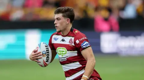 Jacob Douglas, while in action for Wigan Warriors, carrying the ball in his right hand looking up while on the move.