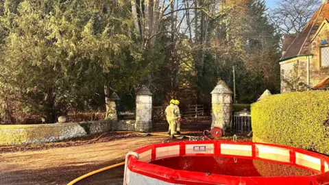 OFRS Two firefighters walking into the property's yard, holding a hose. There is rubber pool filled with water in front. It is daytime.