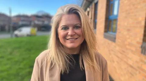 Verity Dean on a residential road, smiling, with a black jumper and camel-coloured coat on.