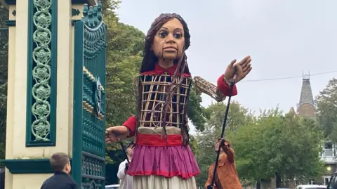 A giant puppet of a girl is walked through the ornate gates of Hull's Pearson Park. She waves at the people welcoming her, including a small boy with his back to the camera. 