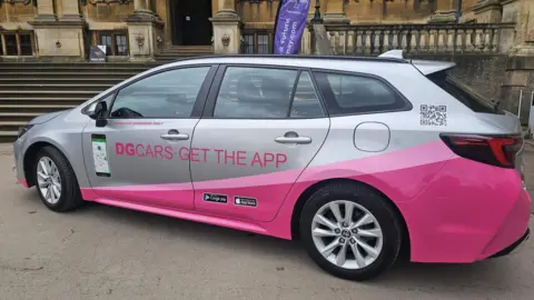 A silver saloon car has a bright pink stripe running from the back of the car to the front wheel.