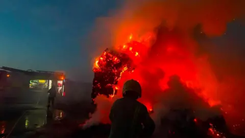 Peterchurch Fire Station A firefighter photographed from behind stands in front of a red burning mound. Red flames are coming off the object with smoke pouring off it.