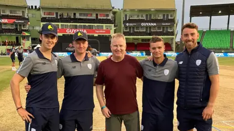 Alan Davis is stood in the middle of four Hampshire cricket players. They are stood in front of a wicket and a large grandstand.