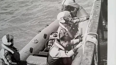 Chris Davey A black and white image of four coastguards on an RNLI lifeboat on water. There is some rope also in shot. 