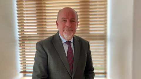 A man in a green tweed jacket is looking directly at the camera. He has very short hair at the sides and has a grey goatee beard. The man, who is also wearing a blue shirt and red tie, is sat in front of a window with horizontal wooden blinds, which are closed.