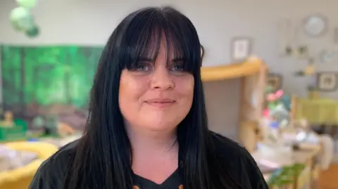A woman in a black t-shirt with long black hair looking at the camera. She is stood in a room with toys behind her. The focus is on her and the room behind is blurred.
