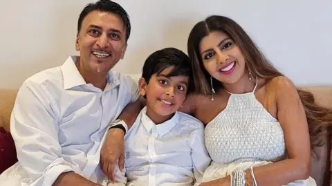 Dad Tarun (left) with a white shirt, dark hair and a moustache,  Arjun (centre) also wearing a white shirt with dark hair and rosy cheeks and Mum Rita (right) in a white halter neck top, wearing pink lipstick and dangly earrings.