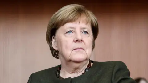 Angela Merkel looks away from the camera, wearing a dark green jacket and brown necklace.