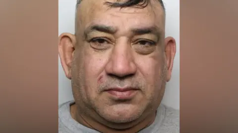 A police mugshot of a man with short black hair wearing a grey prison top