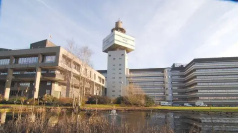 BT Group Part of Adastral Park on a sunny day. A large tower stands between several other office buildings. In front of all the buildings is a large pond. 