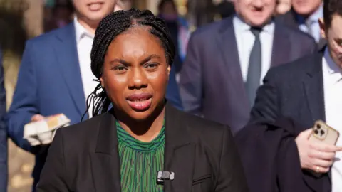 Kemi Badenoch, walking in a crowd of her advisers, on a sunny day in Westminster. She is wearing a green blouse and black jacket. A man behind her is carrying a carton of eggs.