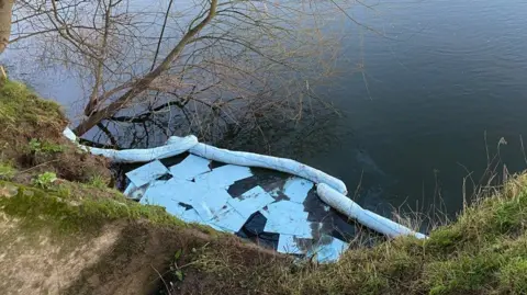 A drainage outlet from the river bank is surrounded by a flexible white boom, with sheets of absorbent fabric inside it on the water's surface. The bare branches of a tree are visible growing from the bank.  