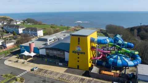 The outside of a water park site, pictured from above. Part of the exterior is bright yellow and reads 'Alpamare'. Water slides can be seen running into the building.