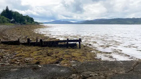BBC A muddy area leads down a gentle slop into a loch with mountains in the background on a cloudy day