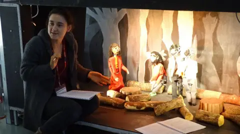 Young People's Puppet Theatre A woman crouching down in front of puppets on a small stage. She has a folder on her lap with lines on it and there is another folder on the stage which has four puppets on it and bits of wood. The woman is wearing a coat which is open and a black top and trousers.