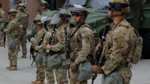 National Guard soldiers, wearing camouflage uniforms and helmets with visors lifted up stationed at the Edward R Roybal federal building in Los Angeles, with more soldiers and police in the background.