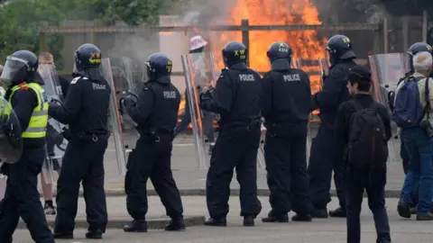 PA Media A line of riot police holding large shields, while a man walks past a generator, with fire burning in the background.