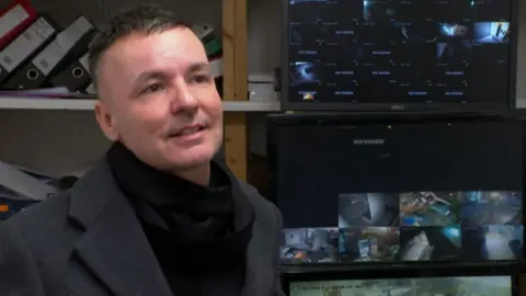 A man with short hair, a black scarf and dark coat, stands in front of a shelving unit which has folders and screens on it.
