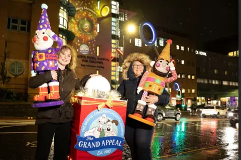 Dani Two ladies are standing at night time in front of the Bristol Children's Hospital beside a large box with the Grand Appeal logo on it of Wallace and Gromit. Both the ladies are smiling to camera and each holding a colourful Aardman Animations Norbot sculpture in their hands. 