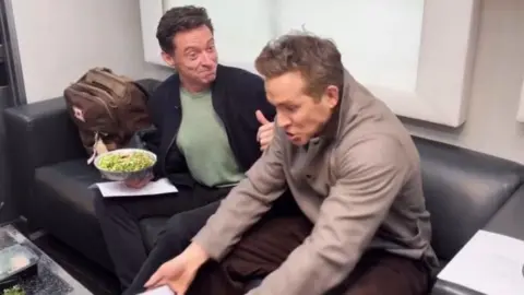 Two men - actors Hugh Jackman and Ryan Reynolds - sit on a sofa. Jackman is giving a thumbs up to Reynolds, who is staring at his laptop, open-mouthed, looking in disbelief 