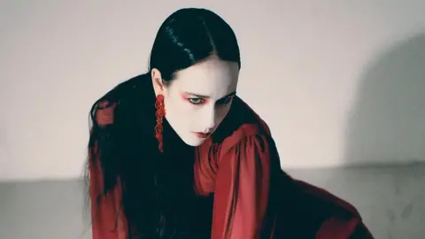 Cal McIntyre Allie X posing in a white room. She is wearing a red dress and long, red earrings. She is staring at the ground and has red makeup around her eyes.