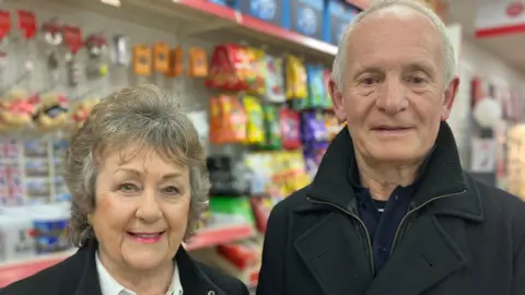 The couple are smiling at the camera and you can see the post office in the background. 