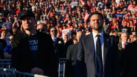 Getty Images Elon Musk and JD Vance look forward, in front of the crowd, with the sun on his face. 