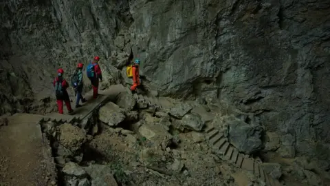 Xiqing Wang / BBC Four people climb down rock stairs into the hole