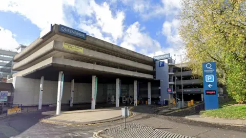 Google the concrete exterior of Queensmere shopping centre multi storey car park. You can see at least four storeys. There are blue signs with Queensmere in white writing. There are several blue P parking signs and a large tree to the right of the car park.