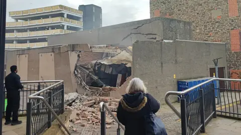 Sam Honeyman A badly damaged wall, with bricks strewn on the ground, following an explosion. Two bystanders survey the scene. 